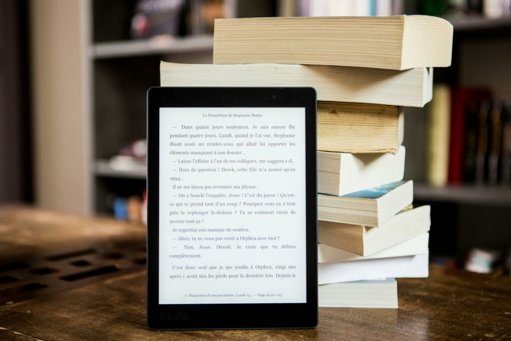 E-reader with text displayed next to a stack of physical books on a wooden table.